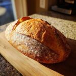 A freshly baked loaf of golden brown Italian bread with a dusted flour crust resting on a wooden cutting board.