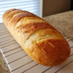 A golden-brown, freshly baked loaf of french bread resting on a white and chrome cooling rack.