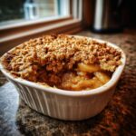 Close-up of a warm, individual serving of pear crisp with a golden oat topping spilling over soft baked pears.