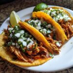 Close-up of three juicy chicken tacos topped with diced white onion and cilantro, served with lime wedges.