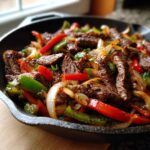 Close-up of sizzling steak fajitas with sliced beef, red and green peppers, and onions in a black cast iron skillet.
