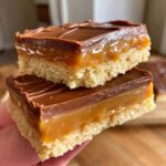 Two stacked, decadent twix cookies bars showing a shortbread base, thick caramel layer, and chocolate topping.