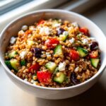 A close-up shot of a vibrant Mediterranean farro salad mixed with cucumber, tomatoes, olives, and feta cheese in a white bowl.