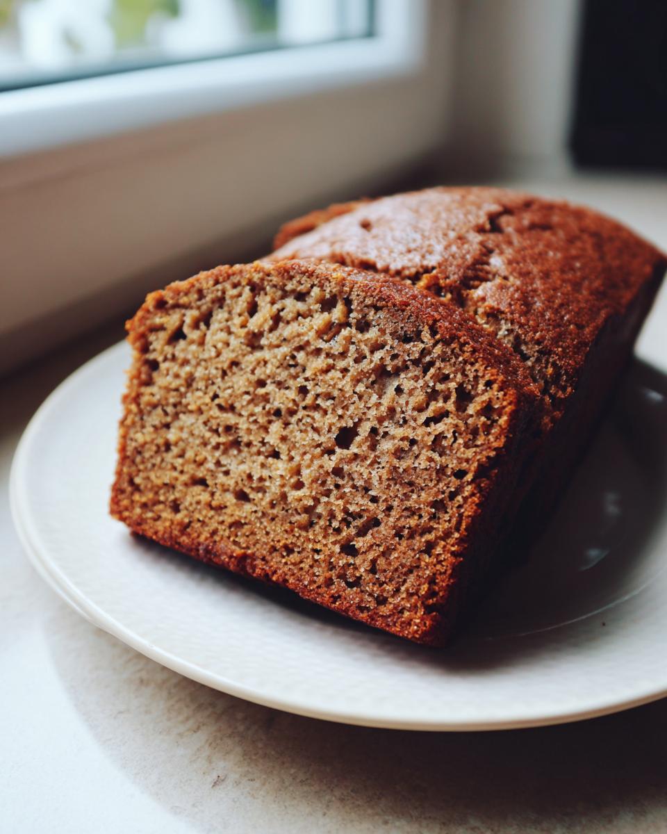 Close-up of a thick slice of moist banana bread recipe loaf on a white plate near a window.