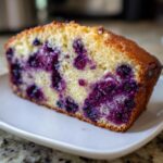 Close-up of a moist slice of blueberry bread showing bright purple blueberries baked into the tender, golden crumb.
