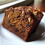 Close-up of two moist slices of dark brown date nut bread studded with walnuts and raisins on a white plate.