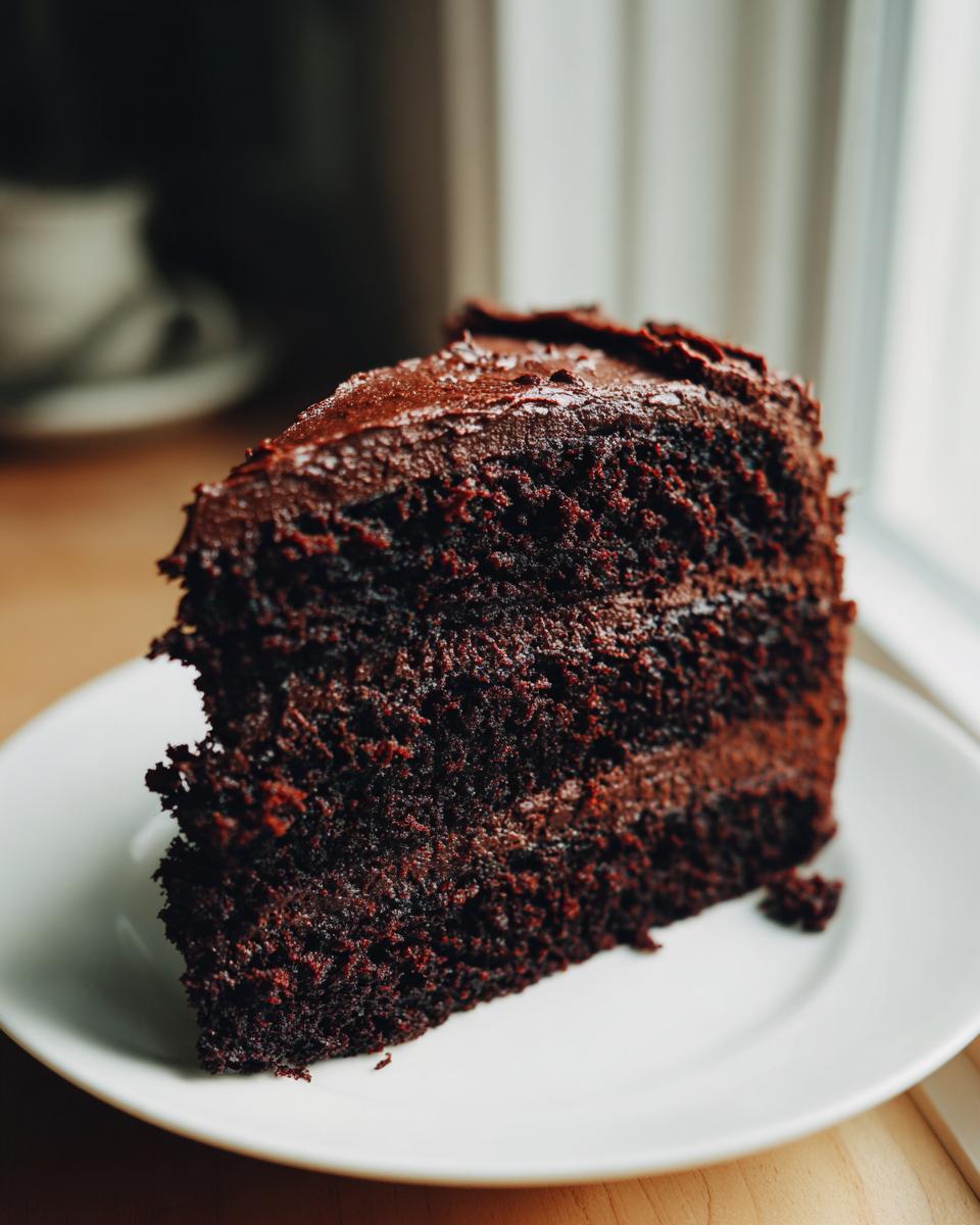 A rich, dark slice of homemade chocolate cake with thick chocolate frosting served on a white plate.