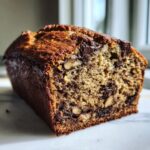 Close-up of a moist vegan banana bread loaf, studded with chocolate chips and walnuts, showing a rich brown crust.