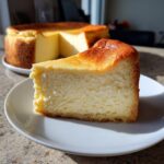 A thick slice of creamy New York Cheesecake with a beautifully browned top sits on a white plate.