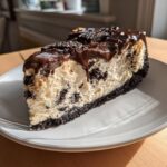 A generous slice of Oreo cheesecake with a dark chocolate crust, creamy filling studded with cookie pieces, and topped with chocolate ganache.