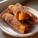 Close-up of crispy, cinnamon-sugar coated peach cobbler egg rolls, one cut open showing the warm peach filling.