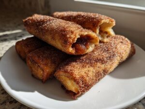 A stack of golden brown peach cobbler egg rolls generously coated in cinnamon sugar, with one broken open showing the filling.