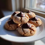 A stack of freshly baked peanut butter blossom cookies, coated in sugar and topped with a chocolate kiss, sitting on a white plate.