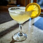 A bright yellow lemon drop cocktail in a coupe glass with a sugar-rim and a backlit lemon slice garnish.