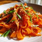 Close-up of a vibrant raw carrot salad featuring wide ribbons of carrot tossed in dressing, topped with sesame seeds and chopped green onions.