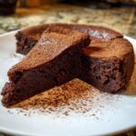 Two slices of dense, dark flourless chocolate cake dusted generously with cocoa powder on a white plate.