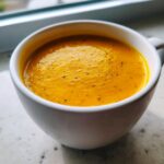 A close-up of vibrant orange roasted butternut squash soup, seasoned with pepper, served in a white mug.
