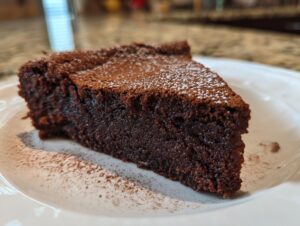 A close-up of a dense slice of flourless chocolate cake dusted with powdered sugar on a white plate.