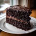 A moist, dark slice of homemade chocolate cake layered with rich chocolate frosting, served on a white plate.