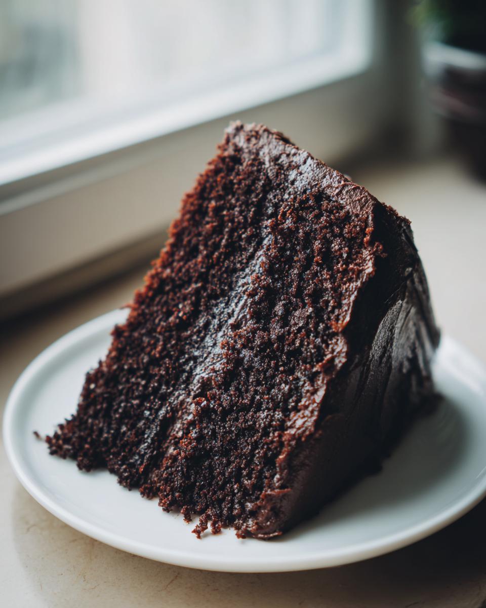 A moist slice of homemade chocolate cake covered in dark chocolate frosting, served on a white plate.
