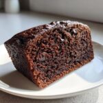 A close-up, sunlit slice of moist chocolate bread with a glossy top, served on a white plate.