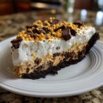 A generous slice of butterfinger pie featuring a dark crust, peanut butter filling, whipped cream, and chopped candy toppings.