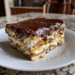 A close-up of a layered slice of no bake eclair cake featuring graham crackers, cream filling, and shiny chocolate frosting.