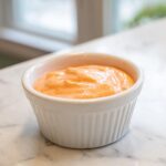 Close-up of creamy, orange-hued smash burger sauce in a small white fluted ramekin.