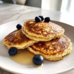 A stack of three golden brown almond flour pancakes topped with fresh blueberries and drizzled with syrup.