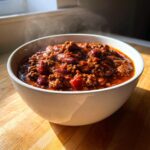 A close-up of a steaming white bowl filled with rich, hearty crockpot chili featuring ground meat and kidney beans.