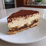 A perfect slice of tiramisu cheesecake showing layers of creamy topping and coffee-soaked base, dusted heavily with cocoa powder.