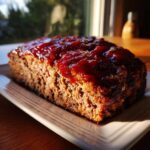 A freshly baked, whole meatloaf topped with a thick, shiny red glaze, resting on a light-colored platter.