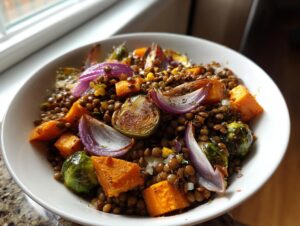 Close-up of a white bowl filled with veggie lentil salad, featuring roasted sweet potato, Brussels sprouts, and red onion.