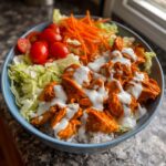 A close-up of a delicious buffalo chicken bowl featuring orange-sauced chicken over rice, topped with ranch dressing, lettuce, carrots, and cherry tomatoes.