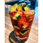 A glass filled with a vibrant red shrimp cocktail mixed with cucumber chunks and topped with sliced avocado.