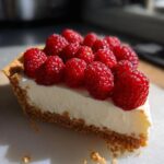 A close-up slice of white chocolate mousse tart with a graham cracker crust, topped generously with fresh red raspberries.